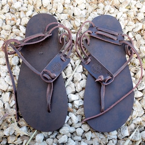 May include: A pair of dark brown leather sandals featuring a flat sole, a thong-style toe post, and thin wrap-around ankle straps. Each sandal includes a stamped circular logo on the upper strap section and the number 44 printed on the footbed.