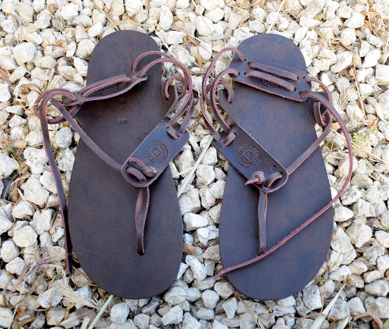 May include: A pair of dark brown leather sandals featuring a flat sole, a thong-style toe post, and thin wrap-around ankle straps. Each sandal includes a stamped circular logo on the upper strap section and the number 44 printed on the footbed.