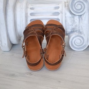 May include: A pair of brown leather sandals with multiple straps and buckle closures. The sandals have a light brown sole and are displayed on a light-colored wooden surface. The sandals are designed for casual wear.