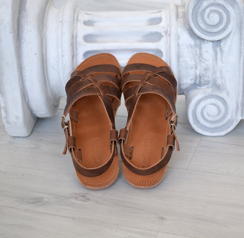 May include: A pair of brown leather sandals with multiple straps and buckle closures. The sandals have a light brown sole and are displayed on a light-colored wooden surface. The sandals are designed for casual wear.