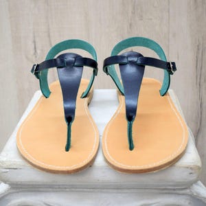 Peut inclure: Paire de sandales en cuir avec un design en T. Les sandales ont une semelle marron clair, une bride bleu marine et une bride de cheville turquoise. Les sandales sont présentées sur une surface blanche sur fond de bois clair.