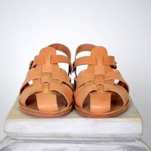 May include: A pair of light brown leather sandals. The sandals have a woven design across the top and a buckle closure on the ankle strap. The sandals are sitting on a white surface.