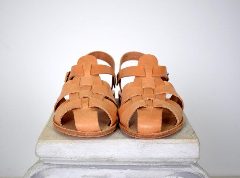 May include: A pair of light brown leather sandals. The sandals have a woven design across the top and a buckle closure on the ankle strap. The sandals are sitting on a white surface.