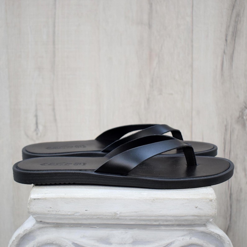May include: A pair of black leather flip-flops. The sandals have a flat sole and a thong strap. The flip-flops are displayed on a white surface against a light wood background. The sandals are a classic design.
