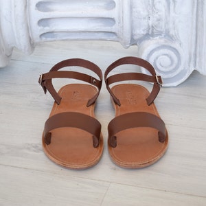 K&ouml;nnte beinhalten: Ein Paar braune Ledersandalen. Die Sandalen haben einen breiten Riemen &uuml;ber den Zehen und einen d&uuml;nnen Kn&ouml;chelriemen mit Schnalle. Die Sohlen sind hellbraun. Perfekt f&uuml;r warme Tage.