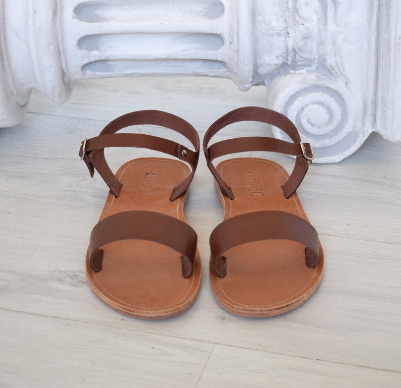 K&ouml;nnte beinhalten: Ein Paar braune Ledersandalen. Die Sandalen haben einen breiten Riemen &uuml;ber den Zehen und einen d&uuml;nnen Kn&ouml;chelriemen mit Schnalle. Die Sohlen sind hellbraun. Perfekt f&uuml;r warme Tage.