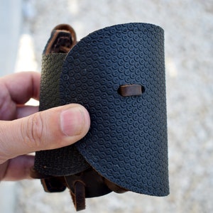 Puede incluir: Un brazalete de cuero negro con un patr&oacute;n circular texturizado. El brazalete tiene un dise&ntilde;o curvo con una correa y hebilla de cuero marr&oacute;n. El brazalete se sostiene en una mano.