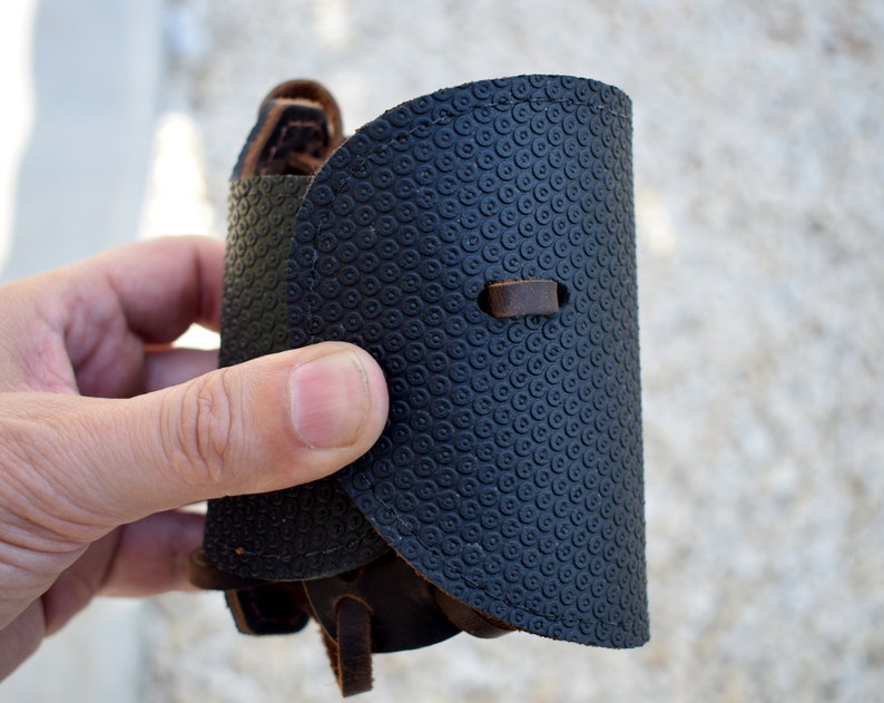 Puede incluir: Un brazalete de cuero negro con un patr&oacute;n circular texturizado. El brazalete tiene un dise&ntilde;o curvo con una correa y hebilla de cuero marr&oacute;n. El brazalete se sostiene en una mano.