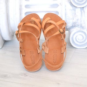 Op de afbeelding: Een paar lichtbruine leren sandalen met meerdere bandjes en gespen. De sandalen hebben een platte zool en een teenlusontwerp. Het leer lijkt glad en de sandalen worden gepresenteerd op een lichtgekleurd oppervlak.