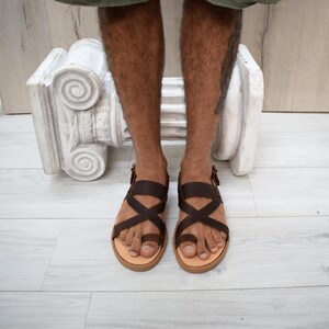 May include: Dark brown leather sandals featuring a toe loop design and crisscross straps across the top of the foot. The sandals have a light tan footbed and a thin buckle closure on the side, providing a secure fit for the wearer's feet.