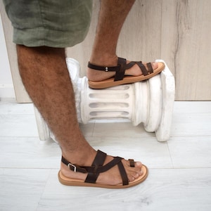 May include: Dark brown leather sandals featuring a toe loop design, multiple thin straps across the foot, and an adjustable ankle strap with a silver-toned metal buckle. The sandals have a flat, light tan sole and a secure heel strap construction.