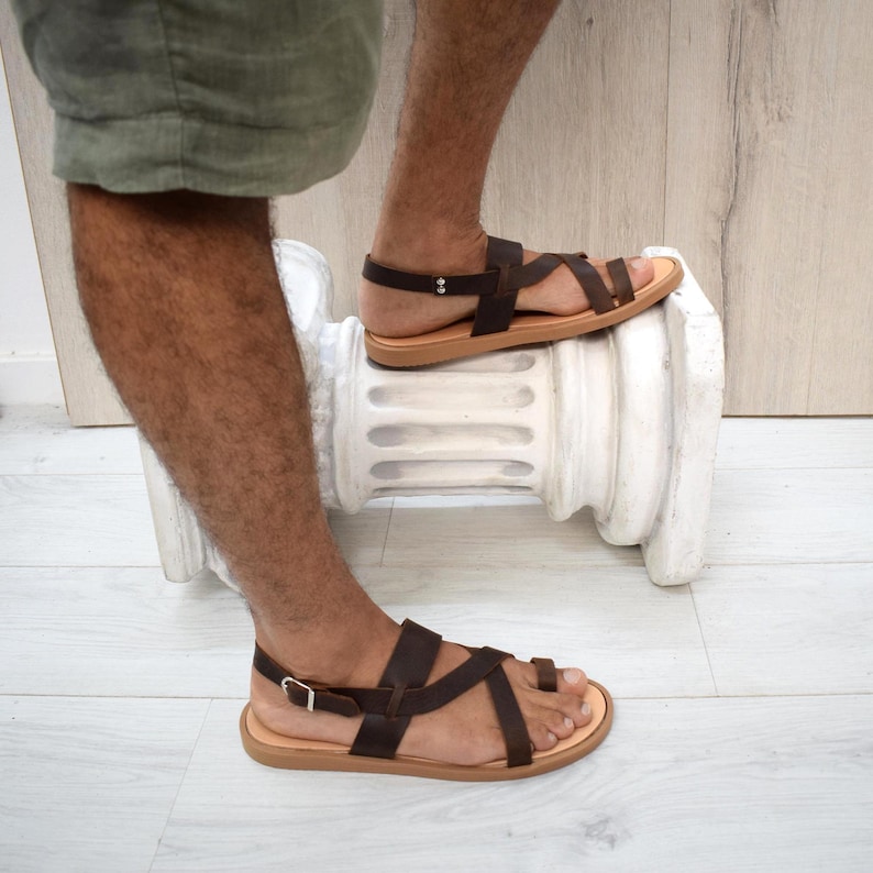 May include: Dark brown leather sandals featuring a toe loop design, multiple thin straps across the foot, and an adjustable ankle strap with a silver-toned metal buckle. The sandals have a flat, light tan sole and a secure heel strap construction.