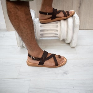 May include: Dark brown leather sandals featuring a toe loop design, multiple crisscrossing straps across the top of the foot, and an adjustable ankle strap with a silver-toned metal buckle. The sandals have a flat, light tan sole.