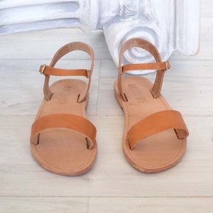 K&ouml;nnte beinhalten: Ein Paar hellbraune Ledersandalen. Jede Sandale hat einen Kn&ouml;chelriemen und einen Riemen &uuml;ber dem Fu&szlig;. Die Sandalen werden auf einer hellen Oberfl&auml;che pr&auml;sentiert. Klassisches Design.