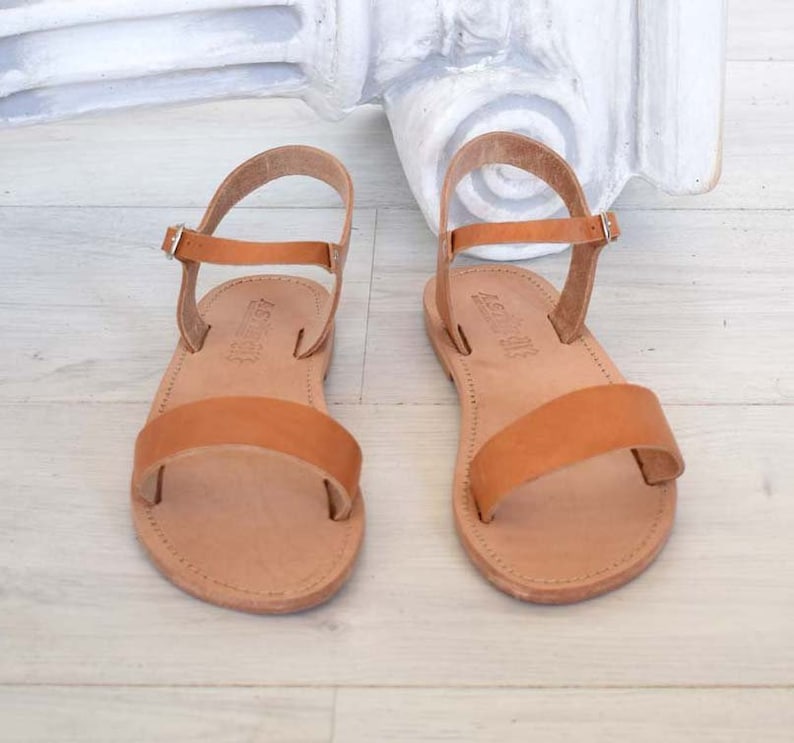 K&ouml;nnte beinhalten: Ein Paar hellbraune Ledersandalen. Jede Sandale hat einen Kn&ouml;chelriemen und einen Riemen &uuml;ber dem Fu&szlig;. Die Sandalen werden auf einer hellen Oberfl&auml;che pr&auml;sentiert. Klassisches Design.