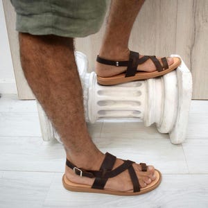 May include: Dark brown leather sandals featuring a multi-strap design with a toe loop, a heel strap, and a silver-toned metal buckle closure. The sandals have a flat, light tan sole and are constructed with thin, adjustable leather straps.
