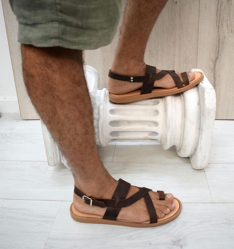 May include: Dark brown leather sandals featuring a multi-strap design with a toe loop, a heel strap, and a silver-toned metal buckle closure. The sandals have a flat, light tan sole and are constructed with thin, adjustable leather straps.