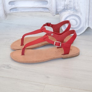 May include: Red leather sandals with a buckle fastening and a thong strap. The sandals have a flat sole and are made of leather.