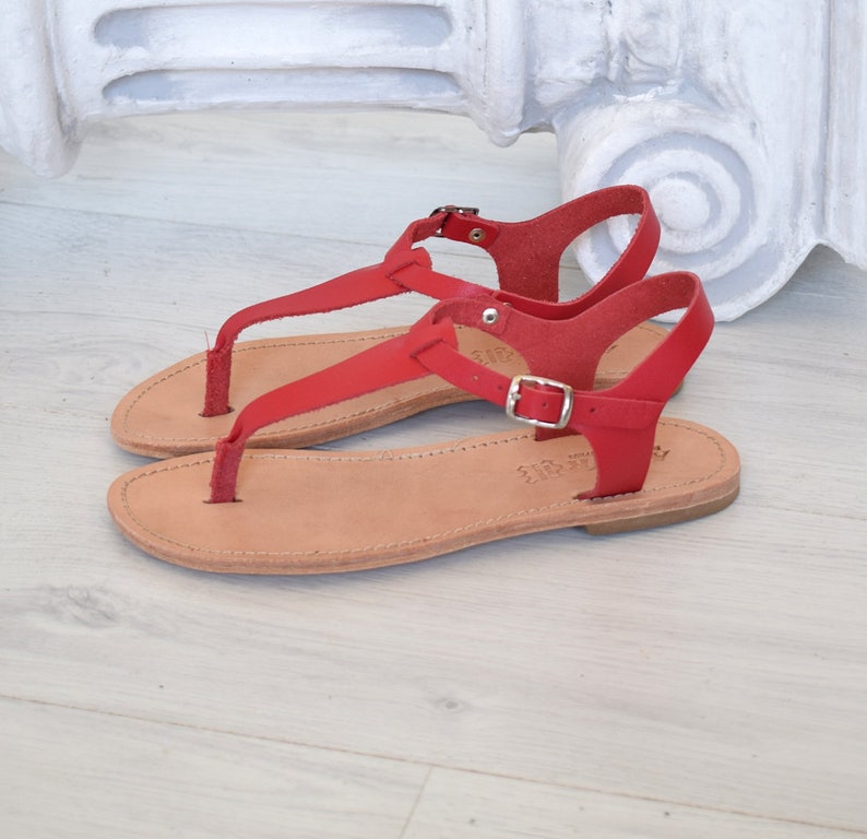 May include: Red leather sandals with a buckle fastening and a thong strap. The sandals have a flat sole and are made of leather.