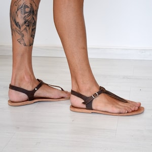 May include: Brown leather sandals with a buckle strap and a thong style toe. The sandals are being worn on a person's feet.