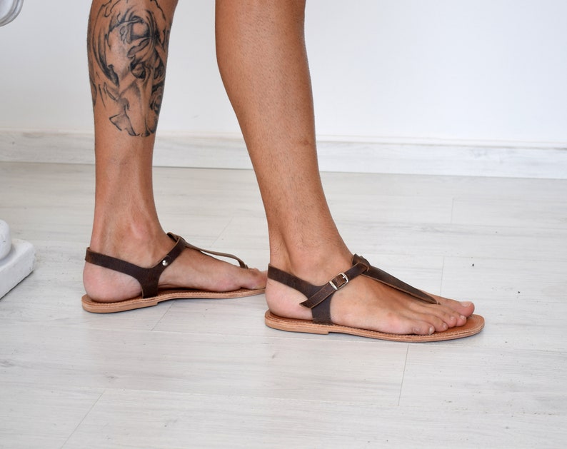 May include: Brown leather sandals with a buckle strap and a thong style toe. The sandals are being worn on a person's feet.