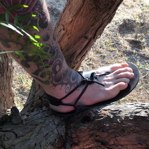 May include: A black leather sandal featuring a minimalist design with thin straps that wrap around the foot and ankle, secured with a simple knot detail at the top of the foot and a flat sole that contours to the shape of the foot.