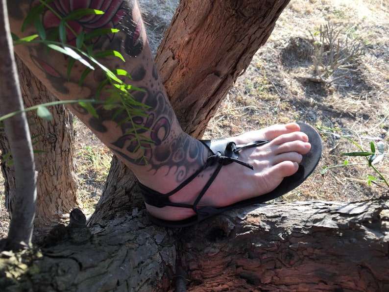 May include: A black leather sandal featuring a minimalist design with thin straps that wrap around the foot and ankle, secured with a simple knot detail at the top of the foot and a flat sole that contours to the shape of the foot.