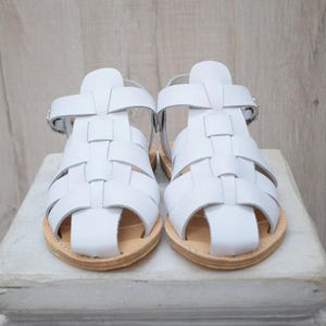 May include: A pair of white leather sandals with a woven design. The sandals have a light brown sole and a buckle closure. The sandals are displayed on a white surface.