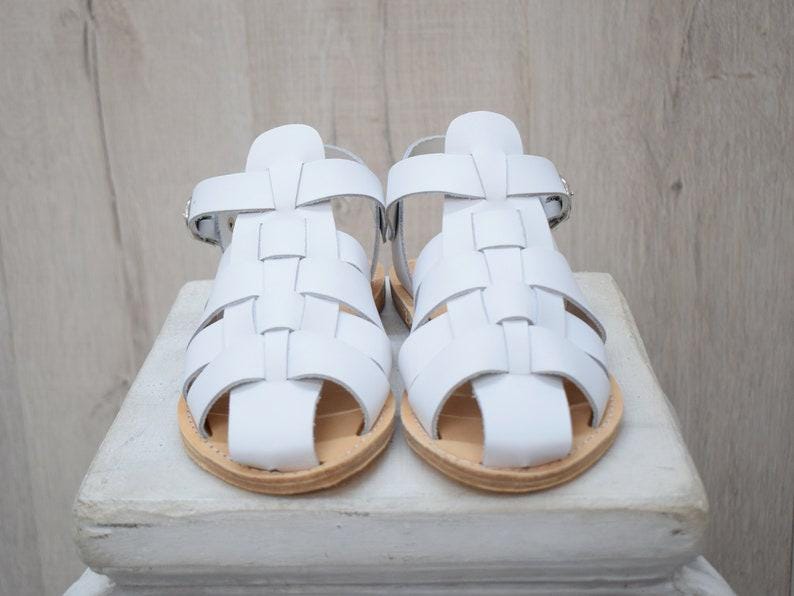 May include: A pair of white leather sandals with a woven design. The sandals have a light brown sole and a buckle closure. The sandals are displayed on a white surface.