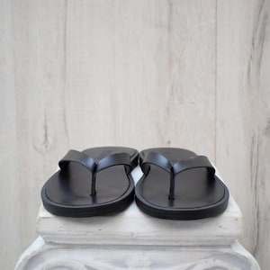 May include: A pair of black leather flip-flops. The sandals feature a simple thong strap design and a rounded sole. The flip-flops are presented on a white surface, set against a light wood-effect backdrop.