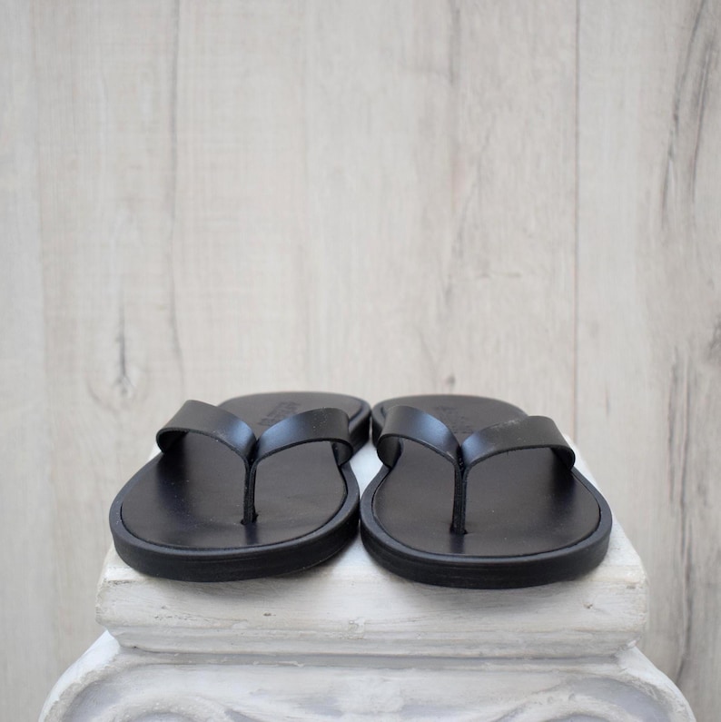May include: A pair of black leather flip-flops. The sandals feature a simple thong strap design and a rounded sole. The flip-flops are presented on a white surface, set against a light wood-effect backdrop.