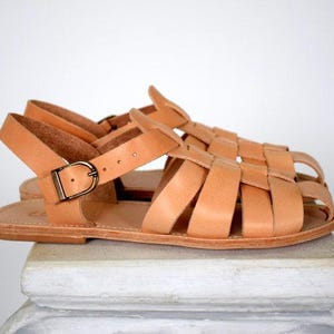 May include: A pair of light brown leather sandals with a woven design and adjustable ankle straps. The sandals have a buckle closure and are displayed on a white surface.