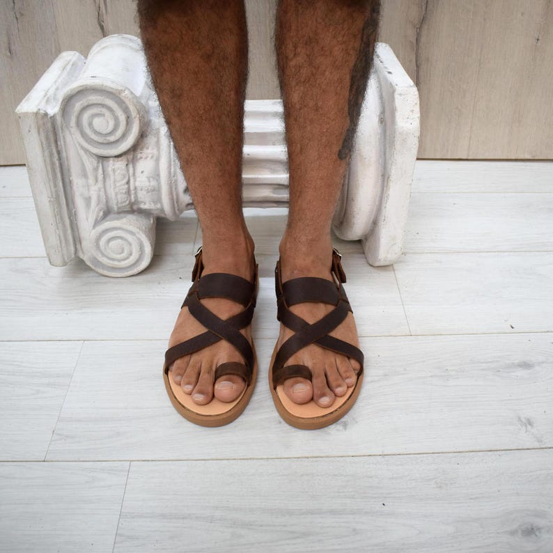 May include: A pair of dark brown leather sandals featuring a toe loop design and crisscross straps across the top of the foot. The sandals have a flat, light-colored sole and a small metal buckle on the side for an adjustable fit around the ankle.