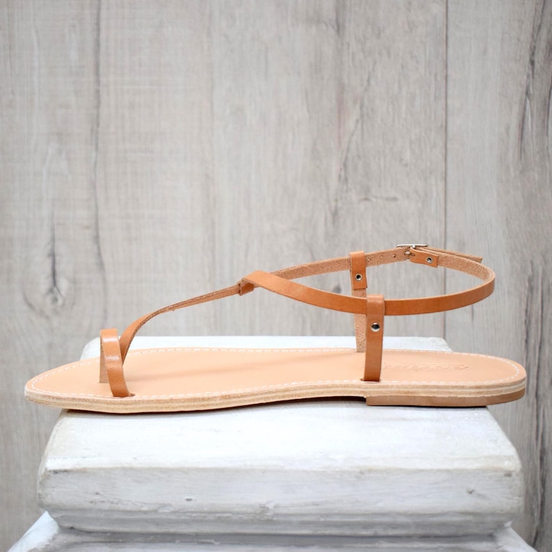 May include: A tan leather flat sandal featuring a thong-style toe post, a thin ankle strap with a small metal buckle, and a flat sole. The sandal has a minimalist construction with slender straps that secure around the foot and ankle.