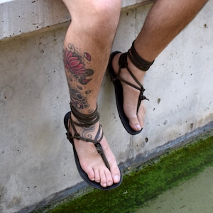 Puede incluir: Sandalias de cuero negro con correas envolventes de color marr&oacute;n oscuro. Las sandalias tienen un dise&ntilde;o sencillo de tanga. Se ve una pierna con un tatuaje de flor de loto.