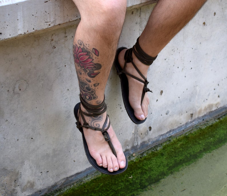 Puede incluir: Sandalias de cuero negro con correas envolventes de color marr&oacute;n oscuro. Las sandalias tienen un dise&ntilde;o sencillo de tanga. Se ve una pierna con un tatuaje de flor de loto.
