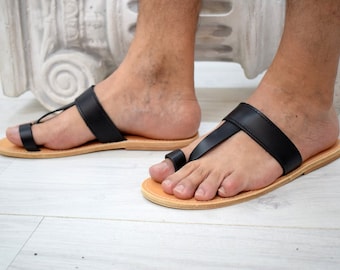 May include: A pair of black leather sandals featuring a flat tan sole and a toe loop design. The sandals have a wide strap across the midfoot that connects to a circular loop around the big toe, providing a simple and open construction for the feet.