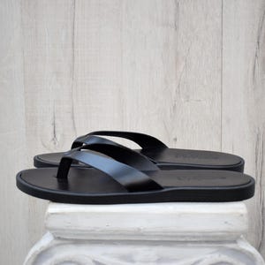 May include: A pair of black leather flip-flops. The sandals feature a simple thong strap design and a flat sole. The flip-flops are displayed on a white surface against a light wood background.