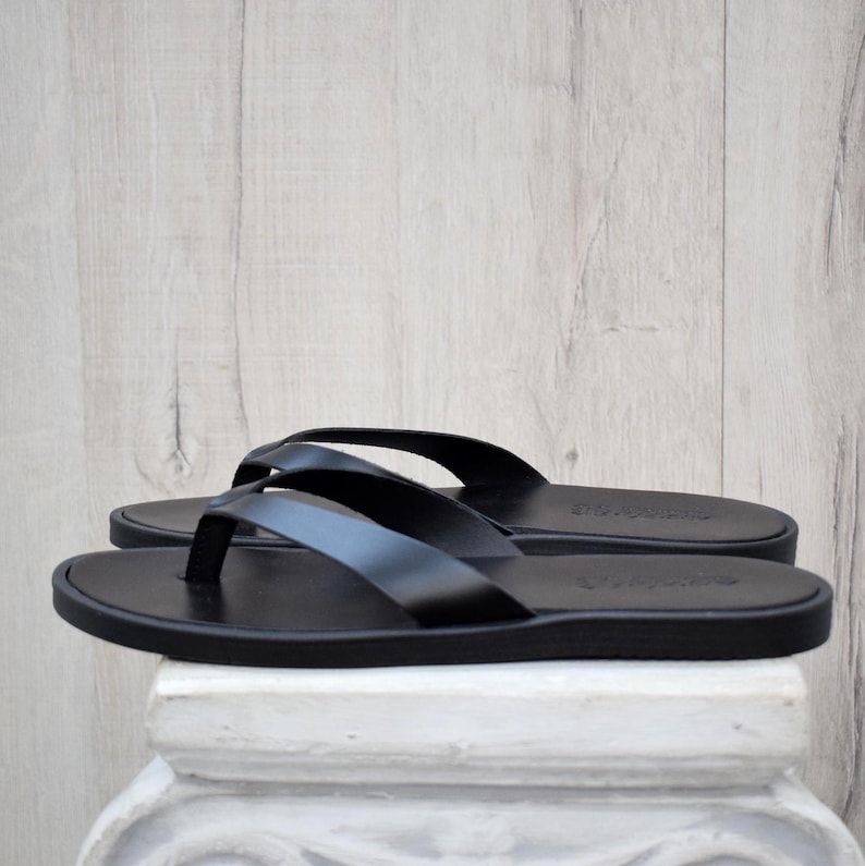 May include: A pair of black leather flip-flops. The sandals feature a simple thong strap design and a flat sole. The flip-flops are displayed on a white surface against a light wood background.