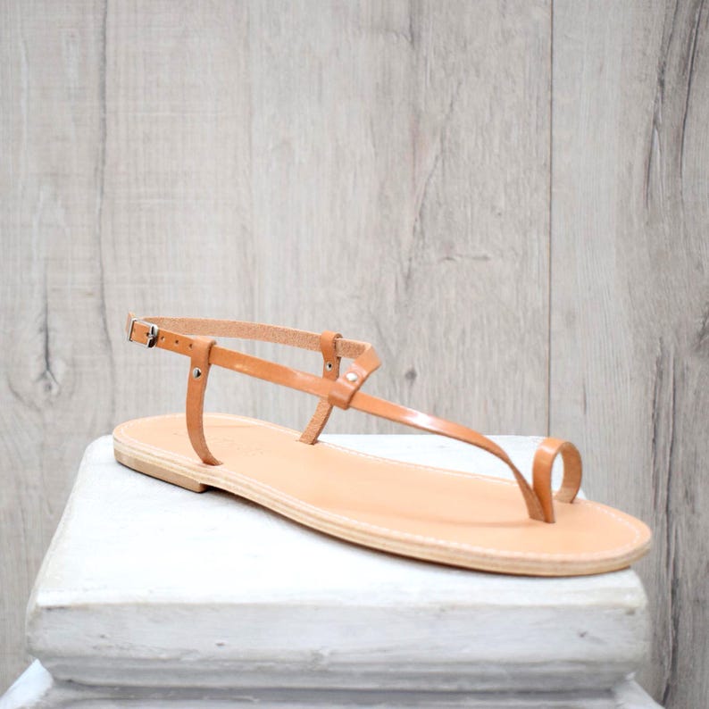 May include: A tan leather flat sandal featuring a minimalist thong-style toe post, a thin ankle strap with a small silver-toned metal buckle, and a flat sole. The design includes a T-strap construction that connects the toe loop to the ankle strap.