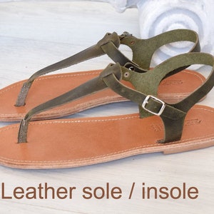 May include: Olive green leather sandals with a brown leather sole and insole. The sandals feature a t-strap design and a buckle fastening.