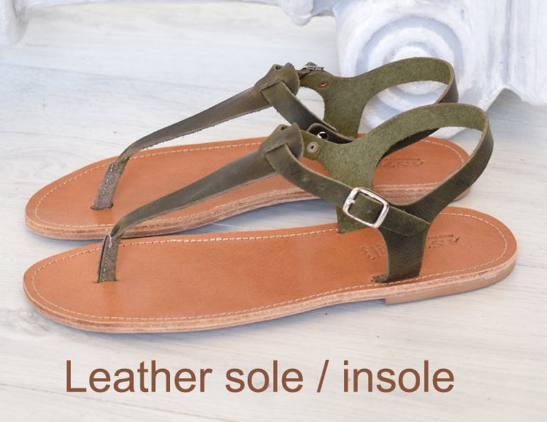 May include: Olive green leather sandals with a brown leather sole and insole. The sandals feature a t-strap design and a buckle fastening.