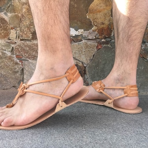 May include: A pair of tan leather sandals featuring a flat sole and a minimalist thong-style design. The footwear includes thin leather straps that wrap around the heel and secure between the toes, showcasing a natural light brown finish throughout.
