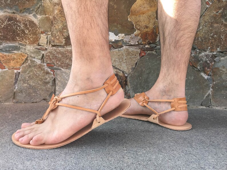 May include: A pair of tan leather sandals featuring a flat sole and a minimalist thong-style design. The footwear includes thin leather straps that wrap around the heel and secure between the toes, showcasing a natural light brown finish throughout.