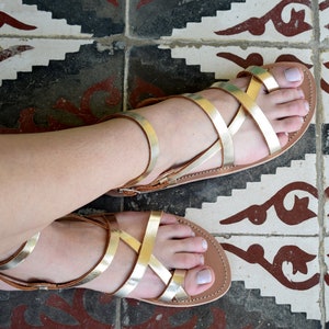 May include: A pair of flat sandals featuring metallic gold straps that crisscross over the top of the foot. The sandals have a tan-coloured footbed and a thin ankle strap with a small metal buckle closure, providing a secure fit for the wearer.