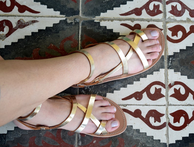 May include: A pair of flat sandals featuring metallic gold straps that crisscross over the top of the foot. The sandals have a tan-coloured footbed and a thin ankle strap with a small metal buckle closure, providing a secure fit for the wearer.