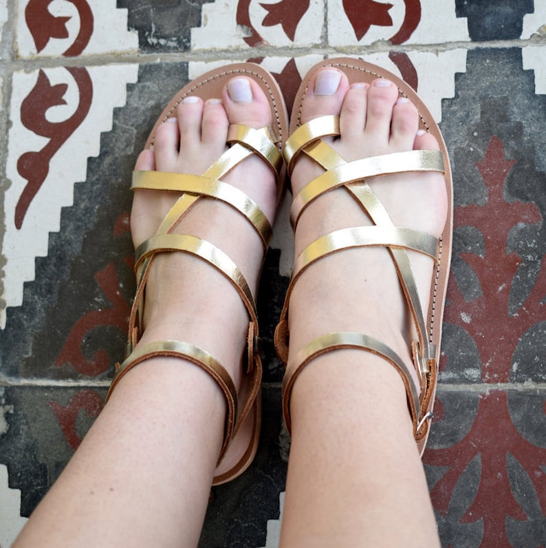 May include: A pair of flat sandals featuring metallic gold straps that crisscross over the top of the foot and include a toe loop. The sandals have a tan leather-coloured sole and a thin ankle strap with a buckle closure on the outer side.