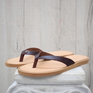 May include: A pair of brown leather flip-flops with light tan soles and brown straps. The sandals are displayed on a white pedestal against a light wood-grain background. The flip-flops are a neutral colour and suitable for casual wear.