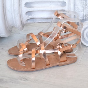 Op de afbeelding: Roze gouden gladiatorsandalen met meerdere banden en een gespsluiting. De sandalen hebben een platte zool en zijn gemaakt van leer.