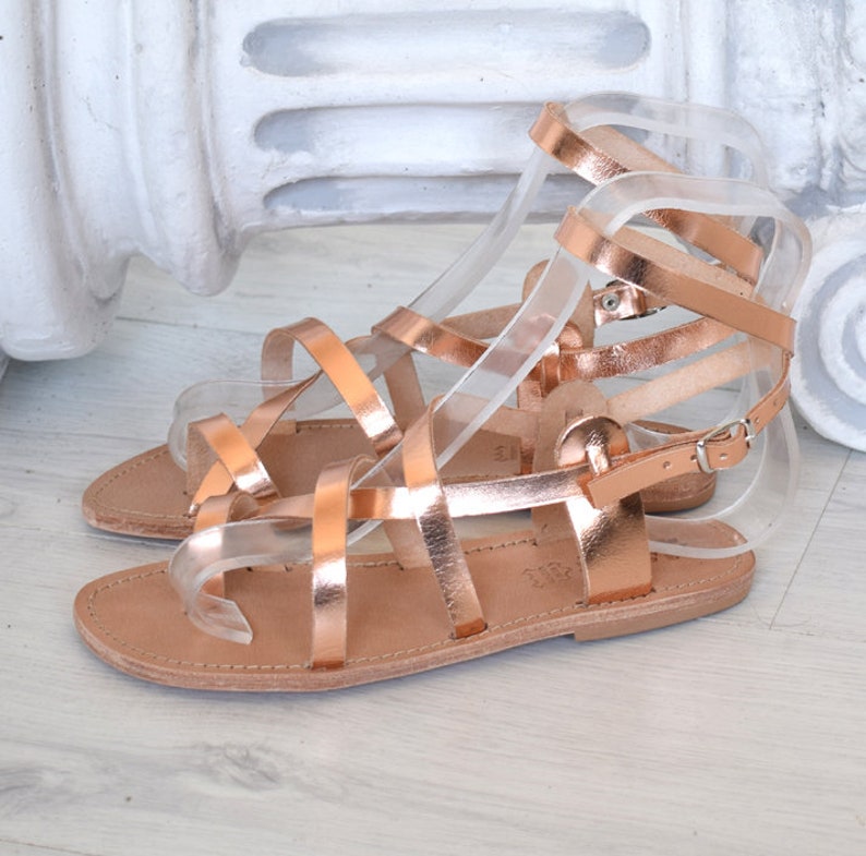 Op de afbeelding: Roze gouden gladiatorsandalen met meerdere banden en een gespsluiting. De sandalen hebben een platte zool en zijn gemaakt van leer.